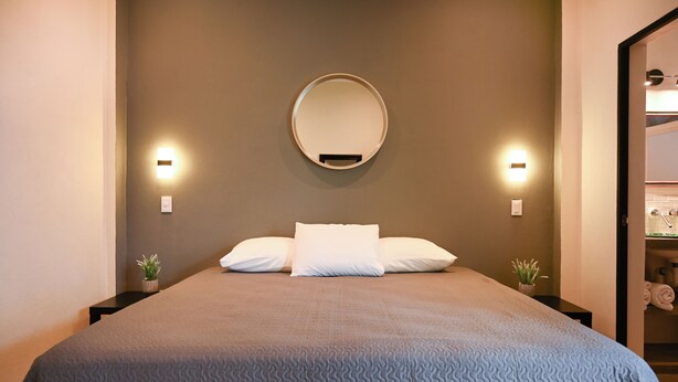 A neatly made bed with a grey textured bedspread, two white pillows, and a round mirror above it. There are two wall-mounted lights on either side of the mirror. - Casa Coral (Sayulita)