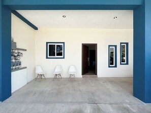 A spacious entryway with a concrete floor, three white chairs, a wall-mounted electrical panel, and two framed pictures.
