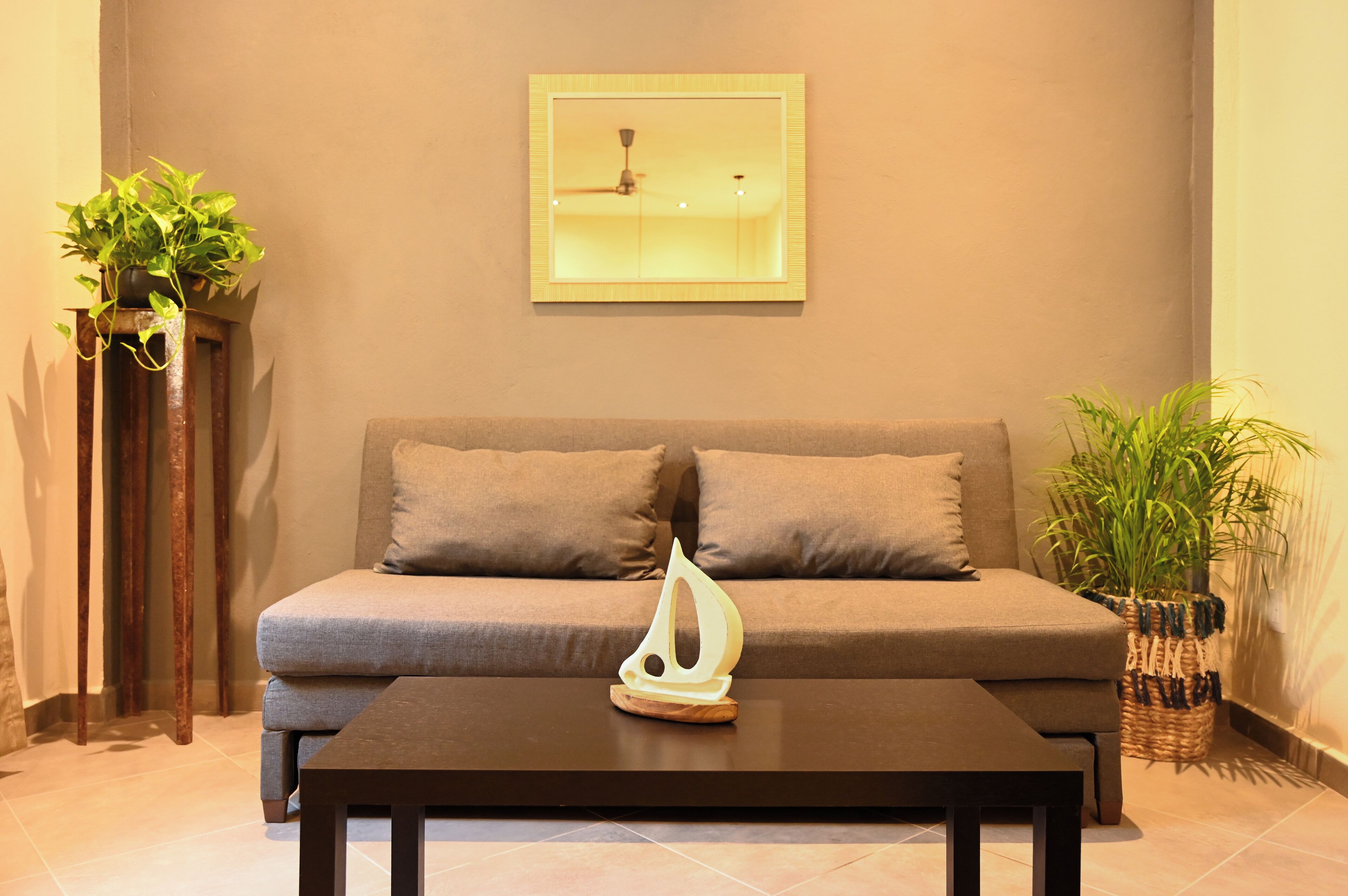 Uma sala de estar com sofá, duas almofadas, mesa de centro com objeto decorativo, ventoinha no teto e dois vasos de plantas.
