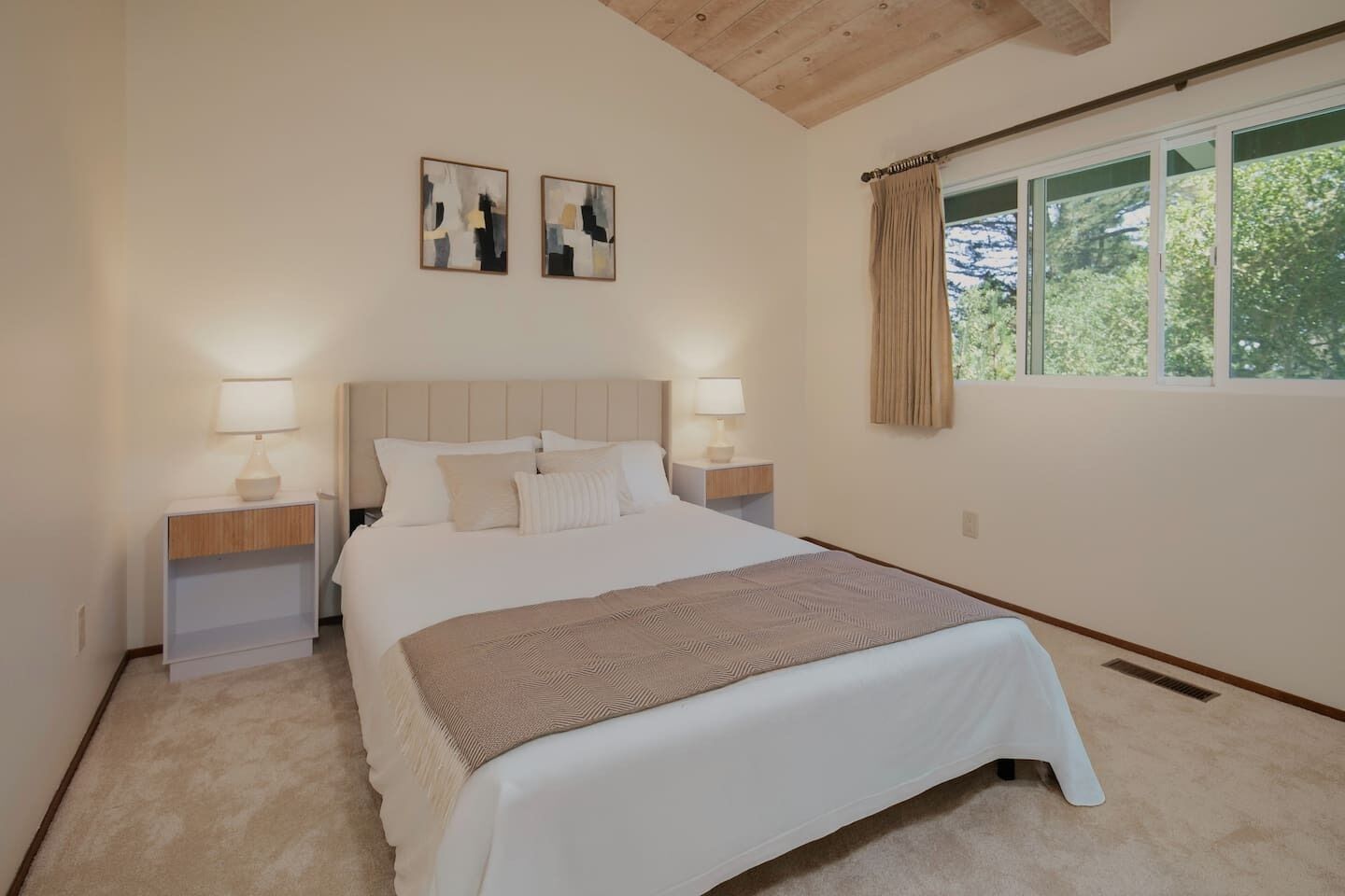 A bedroom with a large bed, two bedside tables with lamps, a headboard, and two abstract paintings on the wall.