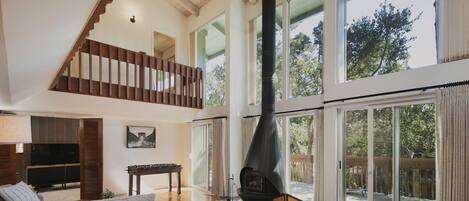 A spacious living room with a high ceiling, a fireplace, a staircase, and large windows offering a view of the outdoors.