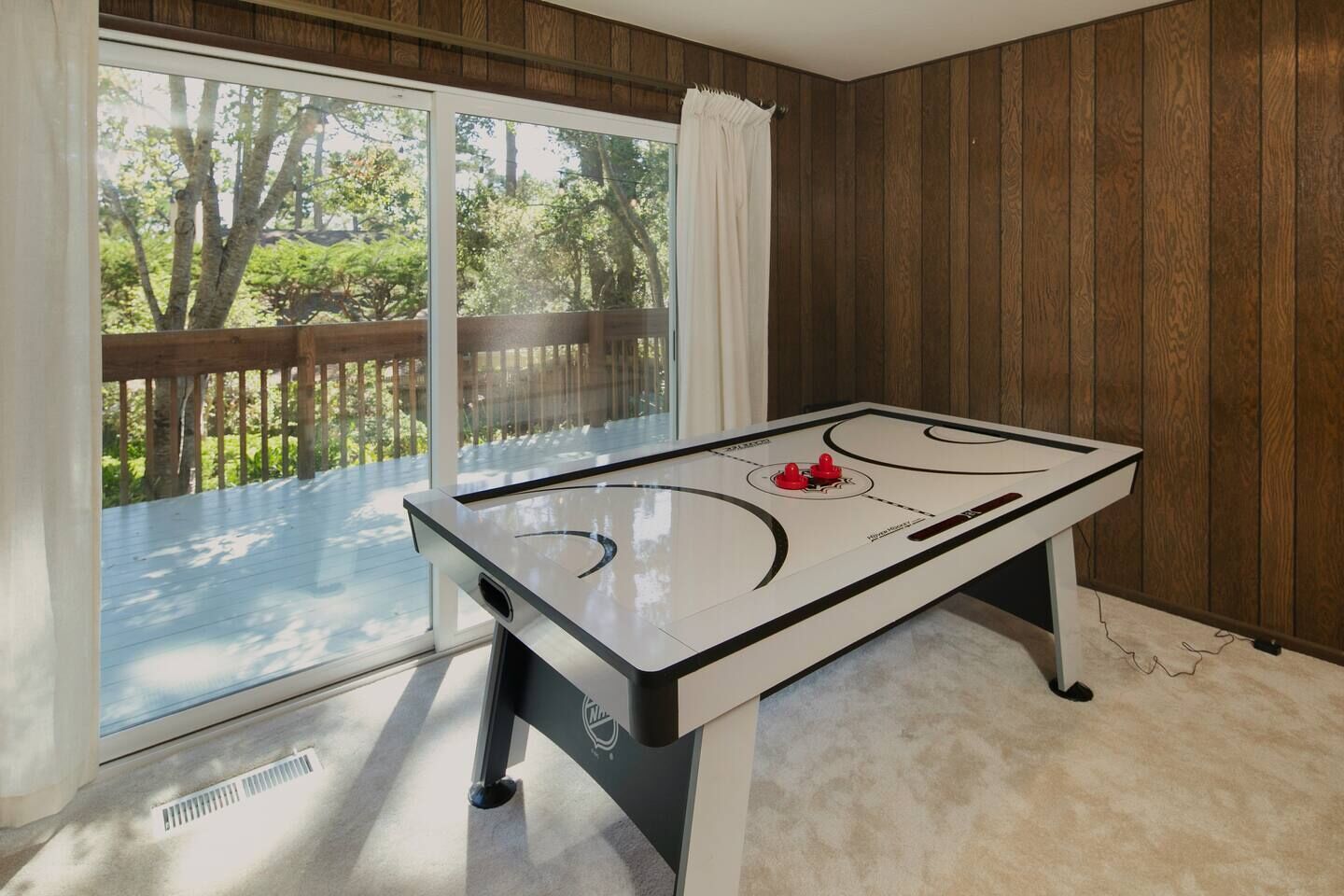A room with a foosball table, large windows, and a view of a wooden deck and trees.