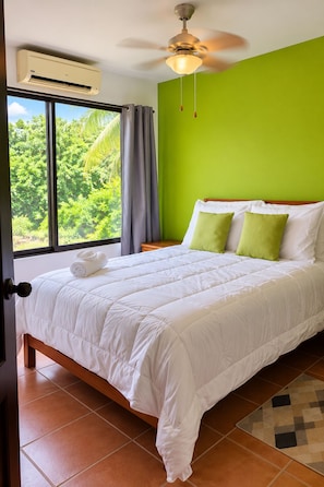 A bedroom with a bed, a ceiling fan, an air conditioning unit, a window with curtains, and a view of greenery outside.