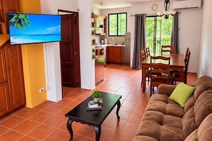A living room with a sofa and a coffee table, a TV mounted on the wall, a dining area with a table and chairs, and a kitchen in the background.