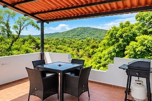 An outdoor patio with a table and chairs, a barbecue grill, and a view of a green landscape.