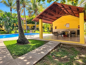 A poolside area with a covered seating area, a pool, and a building in the background.