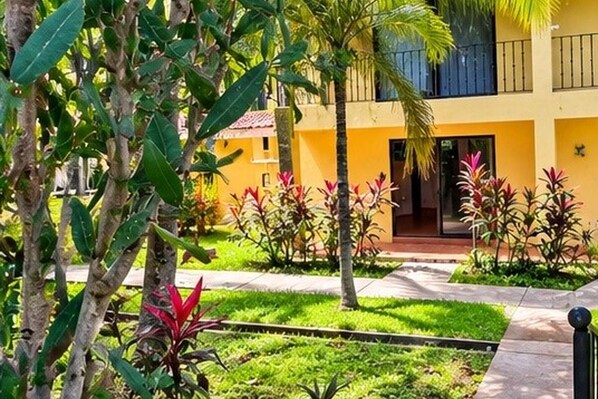 A yellow building with a balcony, surrounded by greenery and plants.