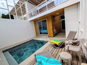 A pool area with lounge chairs and a table, a wooden deck, and a building with a balcony.
