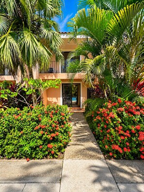 A pathway leading to a building with a pool, surrounded by lush greenery and palm trees.