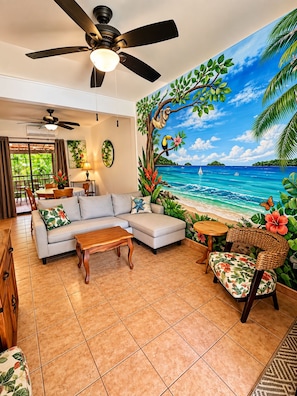 A living room with a sofa, coffee table, and a chair, a ceiling fan, and a mural of a beach scene.