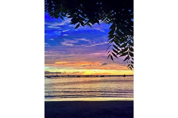 A sunset over a calm sea with boats in the distance and a silhouette of a tree branch in the foreground.