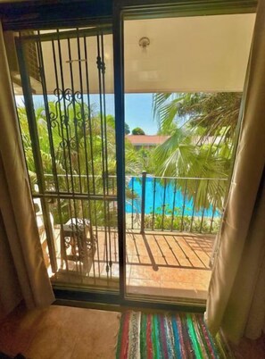 Una habitación con puerta corrediza de vidrio que da a un balcón con piscina, palmeras y suelo de baldosas.
