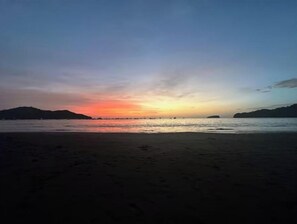 Una playa con un atardecer sobre el agua, siluetas de colinas al fondo y huellas en la arena.
