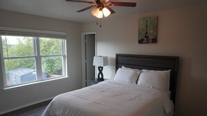 A bedroom with a large bed, a ceiling fan, a window with blinds, and a painting of the Eiffel Tower. - Peaceful Family Retreat – Minutes to AT&T & Rangers Stadiums, Six Flags & More! (Arlington)