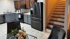 A modern kitchen with grey cabinets, a stainless steel refrigerator, and a microwave. There is a dining area with a table and chairs, and a staircase leading to an upper level. - Peaceful Family Retreat – Minutes to AT&T & Rangers Stadiums, Six Flags & More! (Arlington)