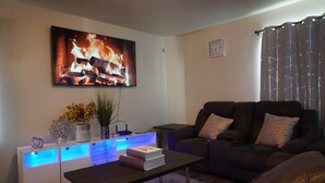 A living room with a flat-screen TV displaying a fireplace, a dark sofa, a coffee table, and a side table with a vase and books. - Peaceful Family Retreat – Minutes to AT&T & Rangers Stadiums, Six Flags & More! (Arlington)