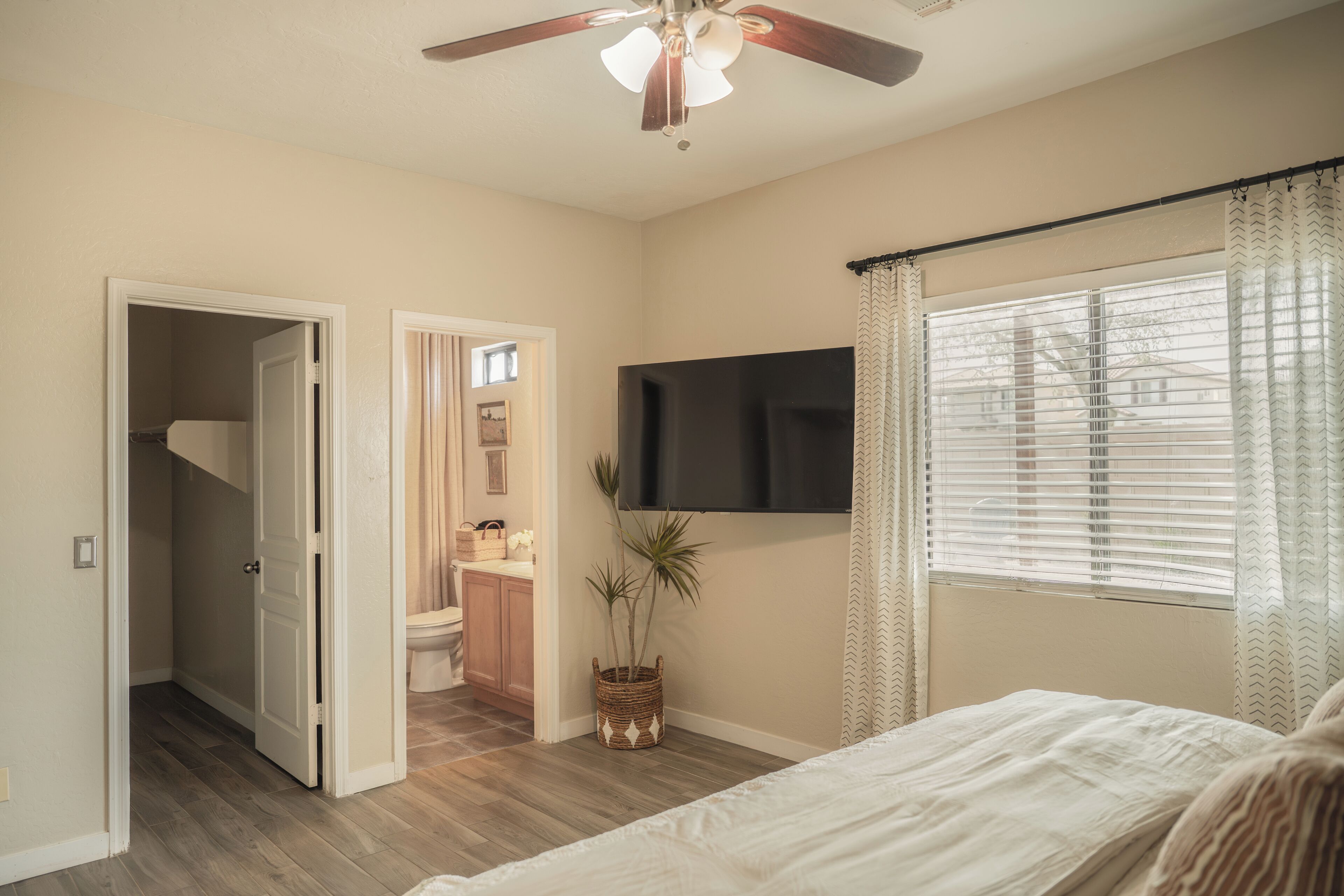 Une chambre à coucher avec un lit, un ventilateur de plafond, un téléviseur fixé au mur et une fenêtre avec des rideaux.