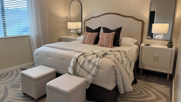 A neatly arranged bedroom with a large bed, two bedside tables with lamps, a mirror, and a small ottoman.
