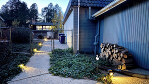 En asfalteret sti der fører til en bygning med garage, omgivet af grøn vegetation og et stabl af brænde.