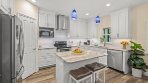 A modern kitchen with white cabinetry, a stainless steel refrigerator, a microwave, and a stove. There are two pendant lights, a sink, and a large island with stools. - Datura Dreams (Sarasota)