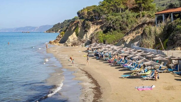 Une plage bordée de rangées de transats et de parasols, une falaise rocheuse parsemée d’arbres, et la mer.