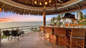 Una terraza con mobiliario de mimbre, una zona de bar y vistas al mar al atardecer.
