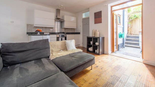 Een moderne woonkamer met een grijze bank, een kleine keuken met witte kastjes, en een houten vloer.