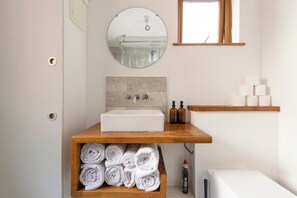 A bathroom with a white sink, a mirror above it, and a wooden shelf below holding rolled towels. - Stratford - Apartment - Sleeps 3 - Great Location (London)