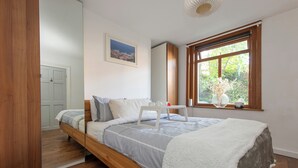 Een moderne slaapkamer met een houten bed, een witte nachtkastje, een raam met uitzicht op groen, en een ingelijst schilderij aan de muur.