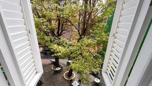 Un área de estar al aire libre con mesas y sillas dispuestas alrededor de un árbol central, delimitada por paredes blancas con contraventanas.