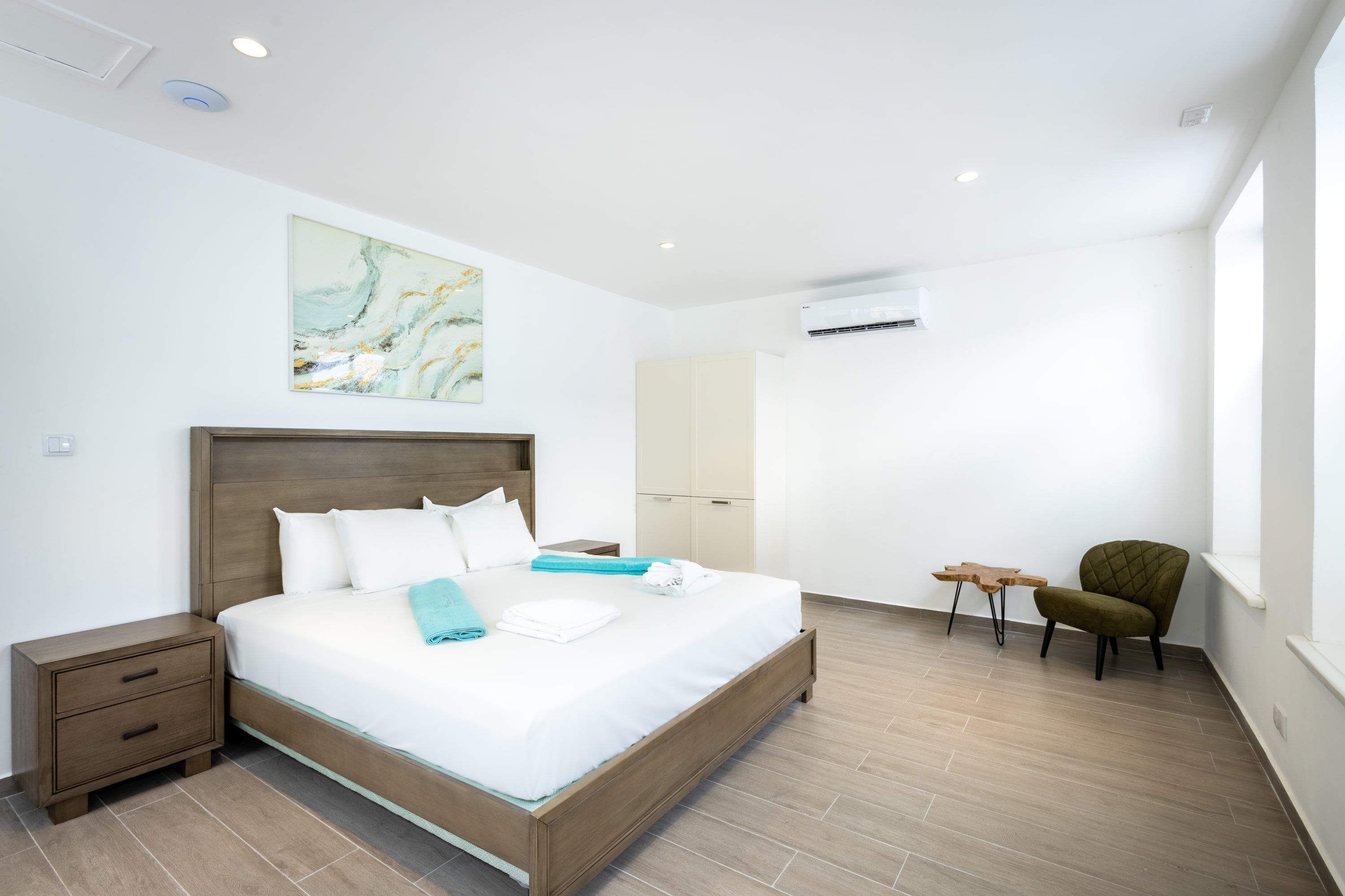 A modern hotel room with a large bed, a nightstand, a small table, a chair, and a painting on the wall.