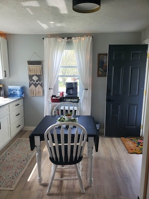 A dining area with a table, chairs, a coffee machine, and a view through a window with curtains. - The Cottage (Burkesville)