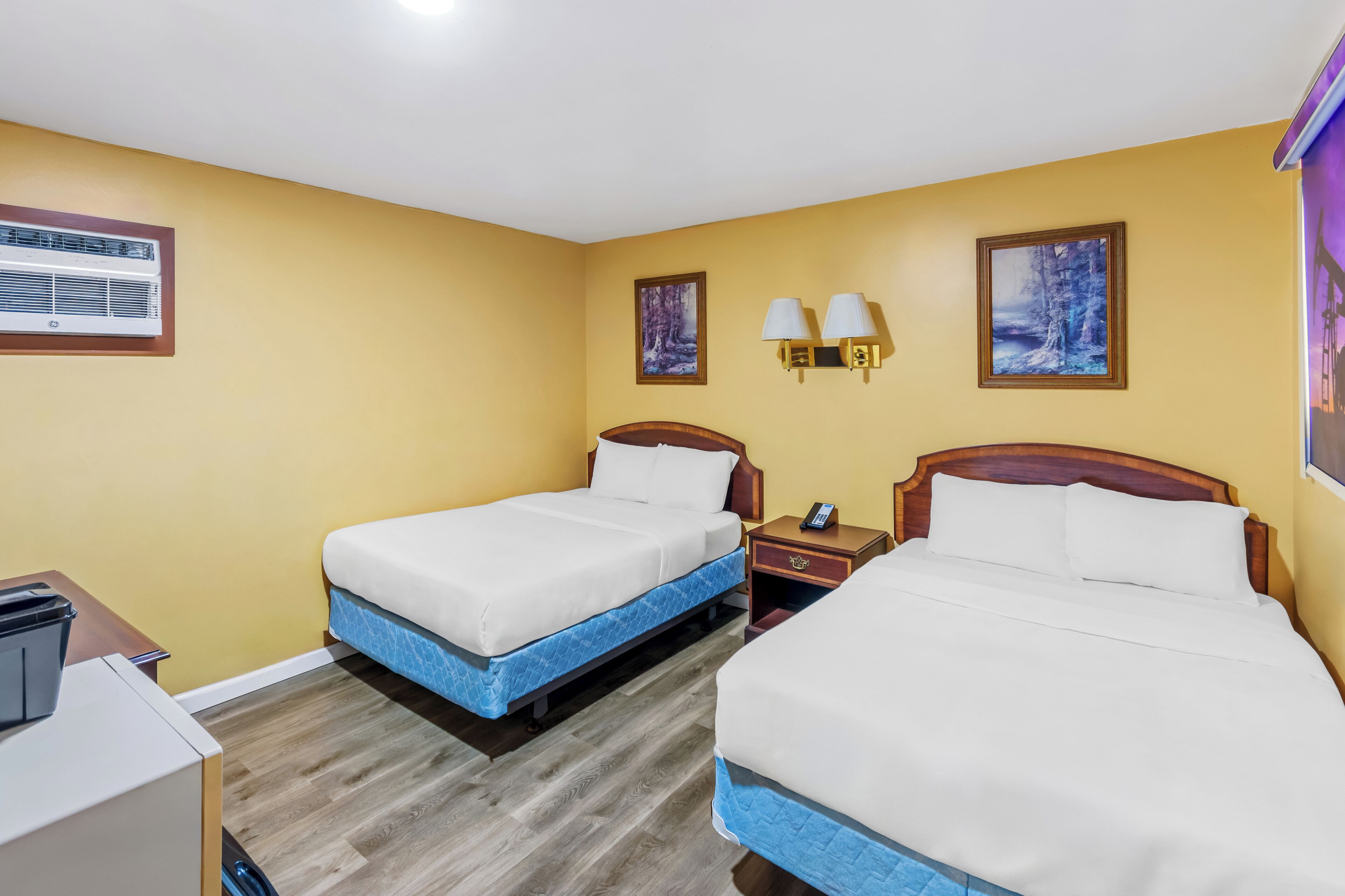 Habitación de hotel con dos camas, un escritorio, una silla, una mesita de noche, una lámpara y aire acondicionado instalado en la pared.