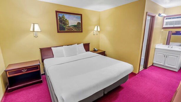 Habitación de hotel con cama, lámparas de noche, mesita de noche, un cuadro, y un baño con lavabo y espejo.