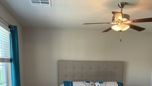 Quarto com uma cama, ventilador de teto, janela com cortinas e uma pequena mesa de cabeceira.