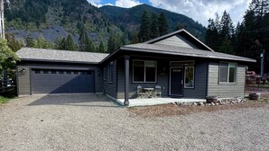 Une maison à deux étages avec un garage, une véranda à l’avant dotée de chaises, et une chaîne de montagnes en arrière-plan.