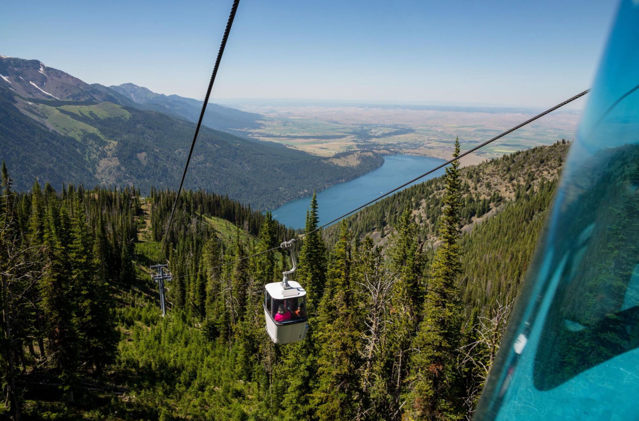 Eine Seilbahn fährt durch eine bewaldete Berglandschaft, im Hintergrund ist ein See zu sehen.