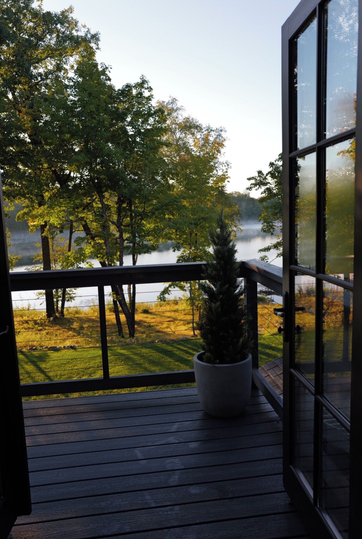 A wooden deck with a potted plant, overlooking a lake with trees and a grassy area.