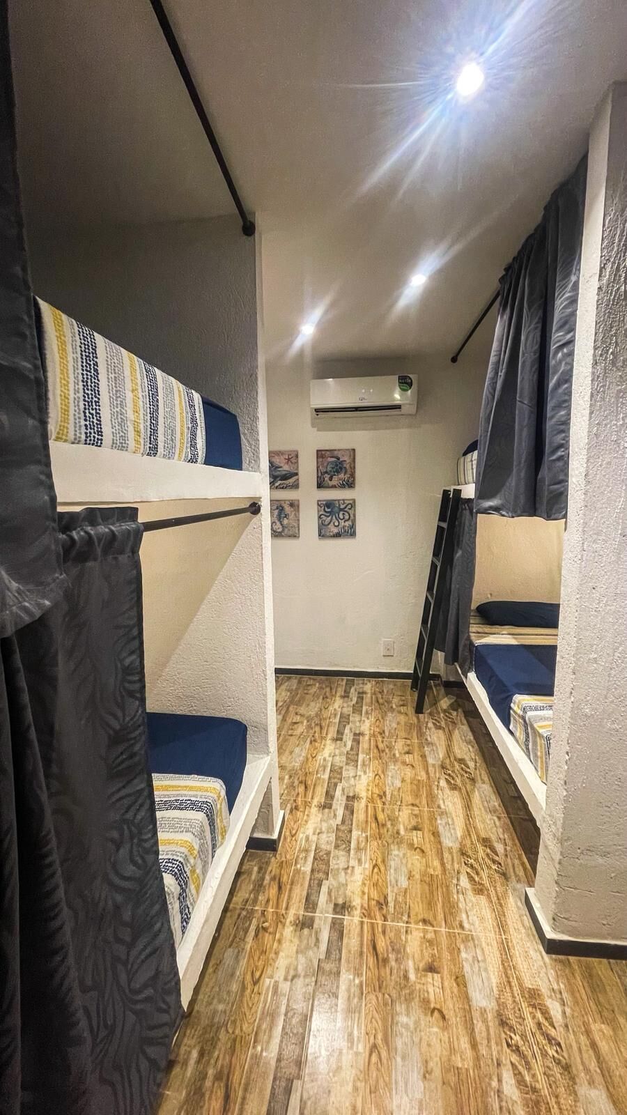 A dormitory room with bunk beds, a wooden floor, an air conditioning unit, and artwork on the walls.