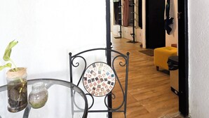 A compact living space with a glass table and chair, a small plant, and a decorative plate. A doorway leads to a hallway with a yellow ottoman and a black bag. - Ocean Lovers Taganga Dive Center& Hostel (Taganga)