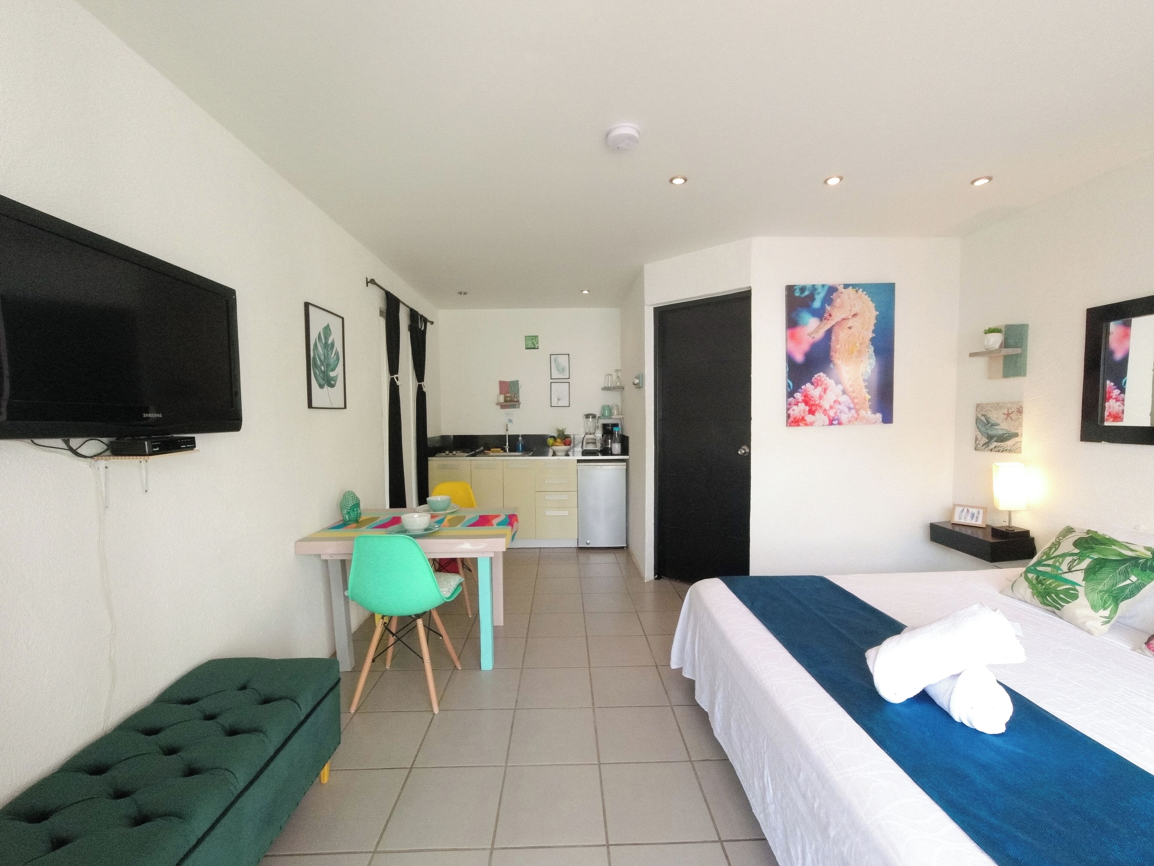 A hotel room with a bed, a desk, a chair, a television, and a kitchenette in the background.