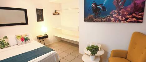 A hotel room with a bed, a green ottoman, a yellow armchair, a small table with a plant, and a painting of an underwater scene.