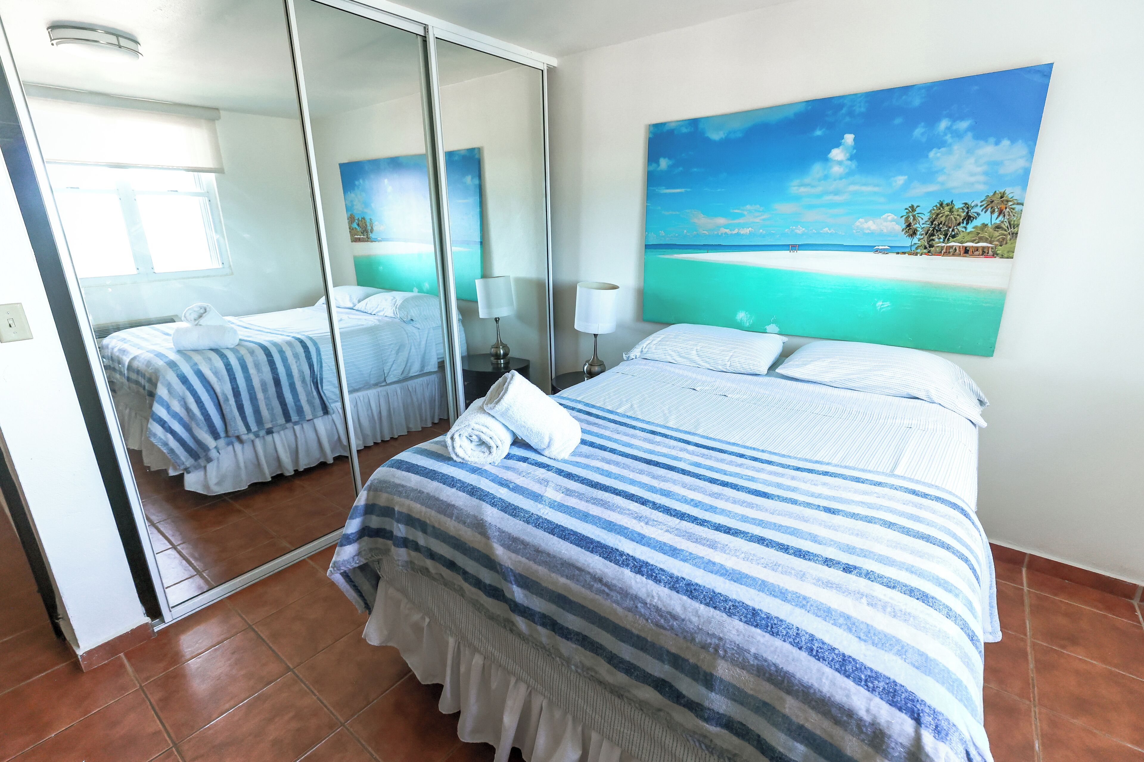 A hotel room with a large bed, a mirror, a painting of a beach, and a window.