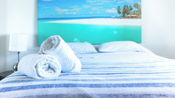 A neatly made bed with a striped blue and white bedspread, a rolled white towel, and a painting of a beach scene on the wall behind. - Coastal Retreat Pool Ocean View Fajardo (Fajardo)
