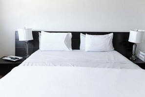 A neatly made bed with white linens, two bedside lamps, and a small table with a remote control and a book.
