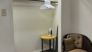 Una habitación compacta con suelo de madera, una pequeña mesa redonda con un hierro, una silla con tapicería estampada y un estante blanco con cestas colgantes.