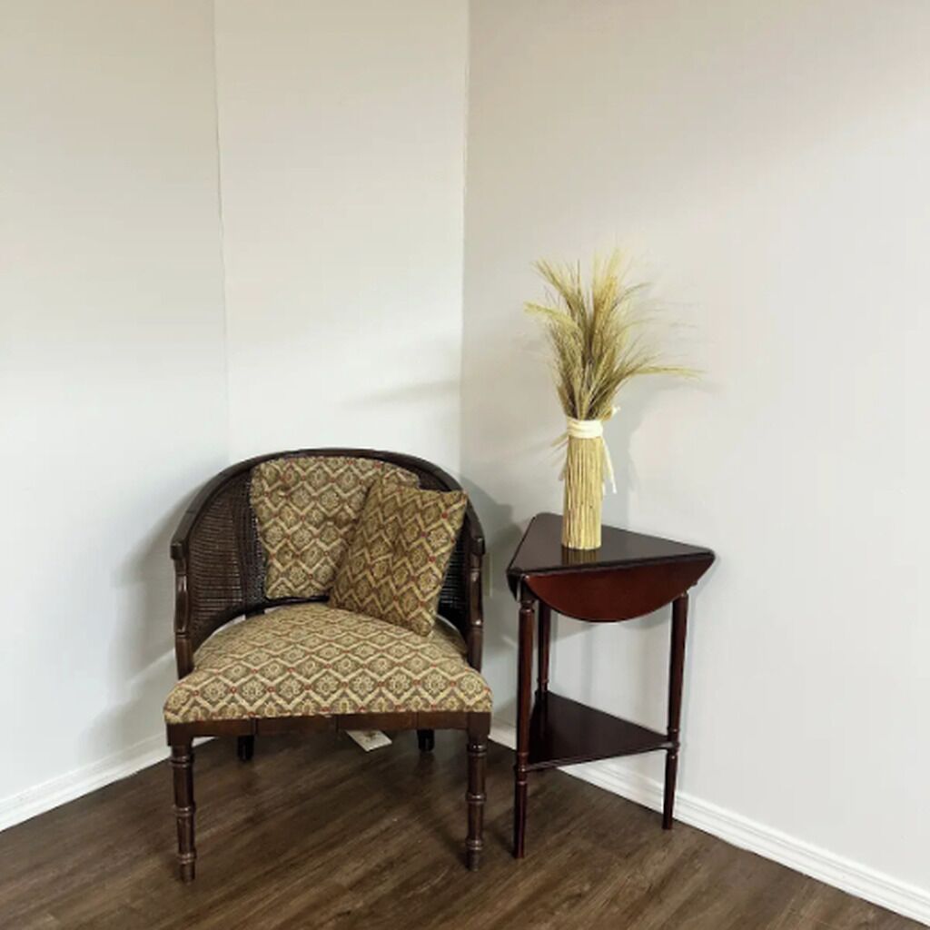A corner room with a dark wood floor, a patterned armchair, and a small table with a vase of dried plants.