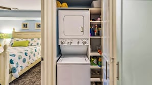 Une petite buanderie équipée d’un lave-linge et d’un sèche-linge, un lit recouvert d’une couvre-lit à motifs floraux et une étagère contenant des produits de nettoyage.