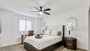 A bedroom with a bed, a desk, a chair, a ceiling fan, and a window with blinds.