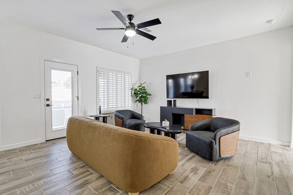 A modern living room with a sofa, two armchairs, a coffee table, a flat-screen TV, a ceiling fan, and a door with a view to the outside.